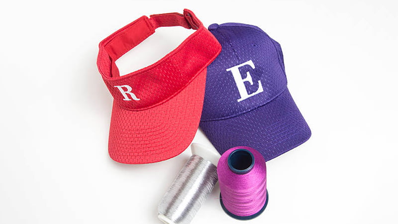 Red visor and purple baseball cap with embroidered letters beside thread spools, showing custom hat embroidery options