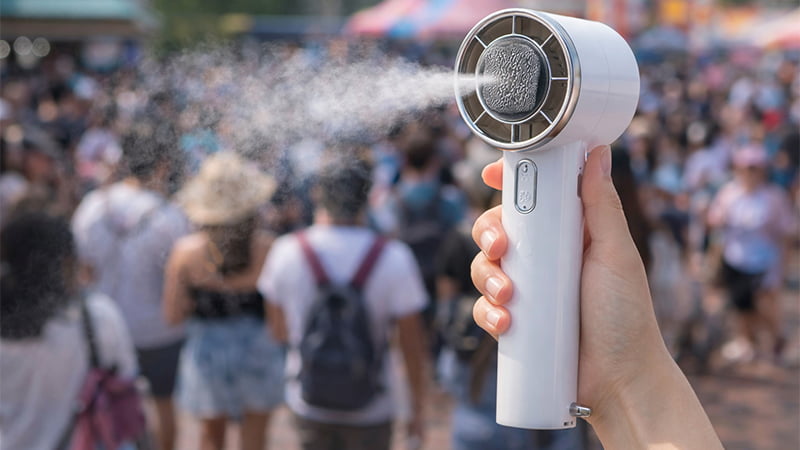Handheld misting fan spraying water mist in a crowded outdoor festival, a cooling summer promotional giveaway.