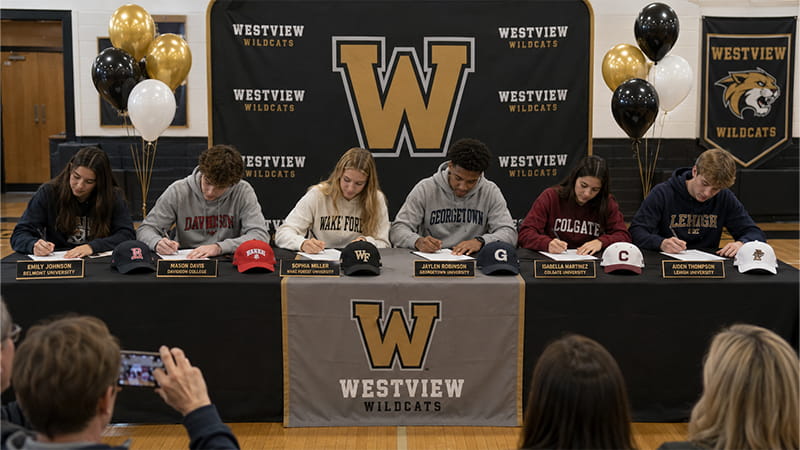 Athletes representing different colleges seated beneath one shared high school backdrop and table banner
